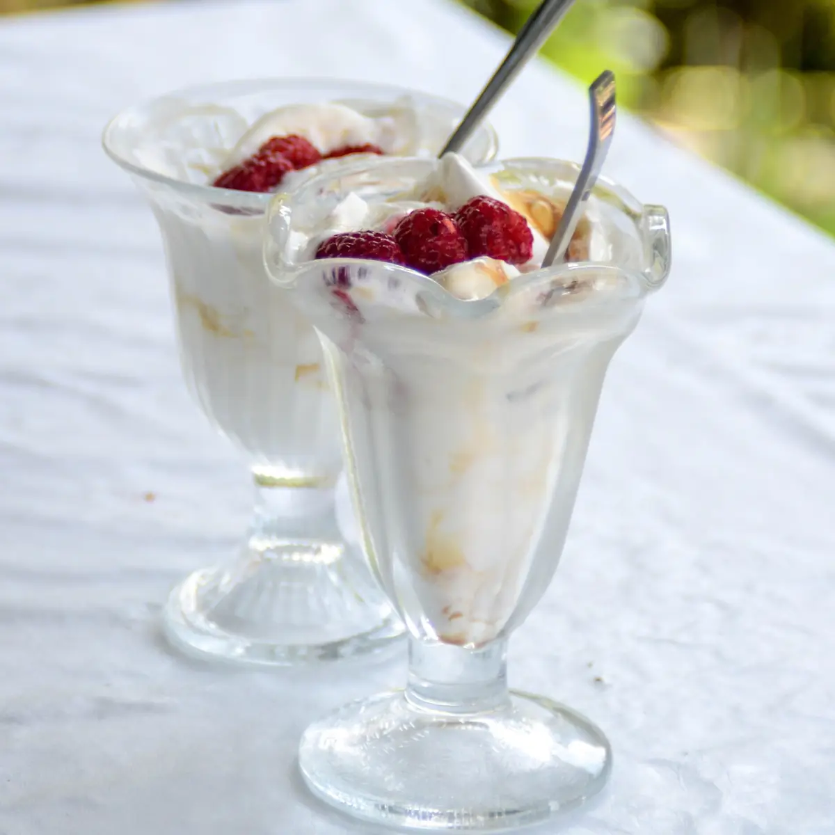 Zwei Eisbecher aus Glas mit hellem Eis und Himbeeren auf einer weißen Tischdecke im Garten
