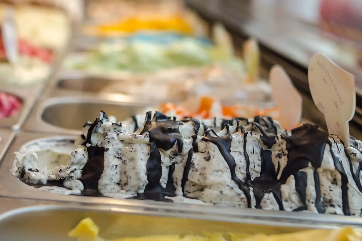 Eistheke mit Stracciatella unter Schokoladenstreifen, dahinter unscharf viele bunte Sorten und Holzschildchen