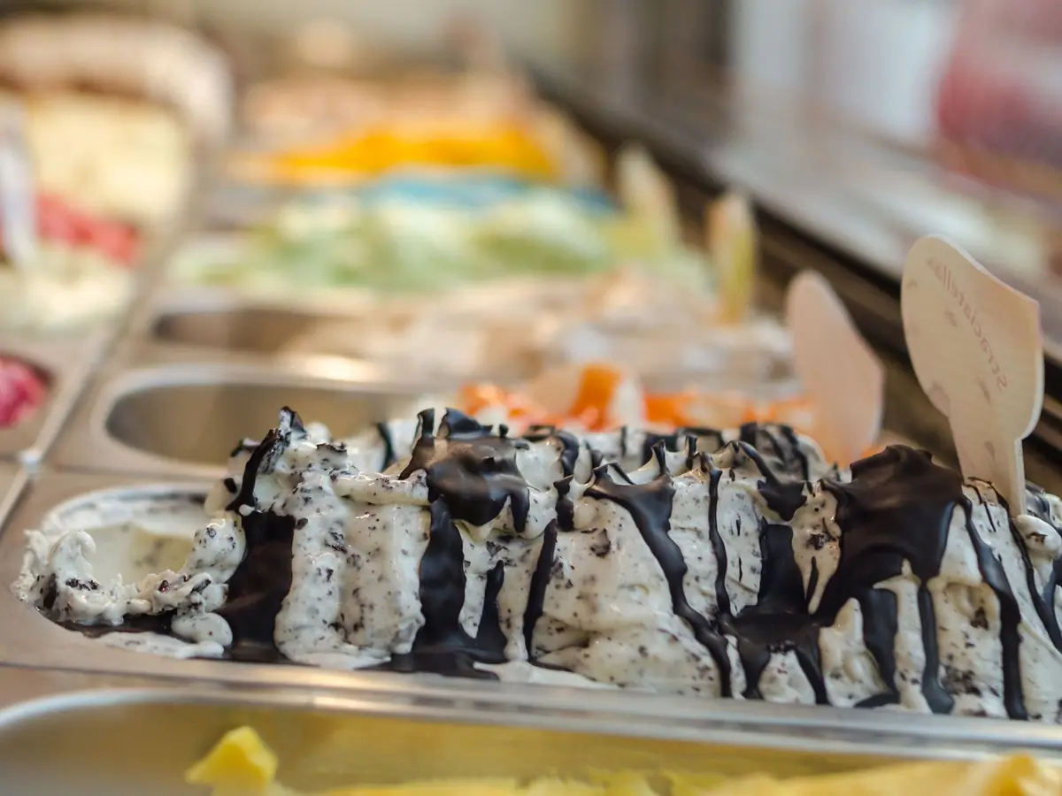 Eistheke mit Stracciatella unter Schokoladenstreifen, dahinter unscharf viele bunte Sorten und Holzschildchen