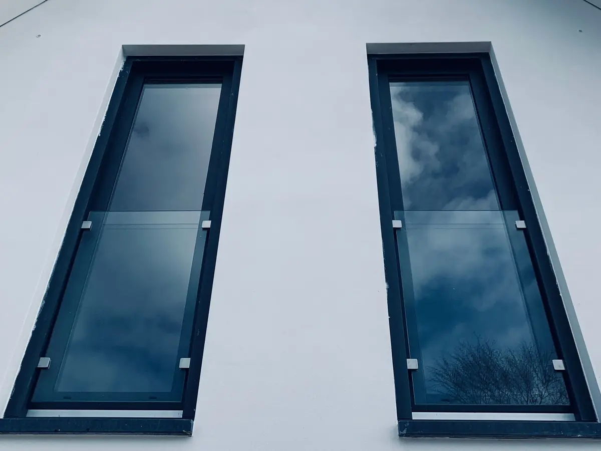 Zwei schmale bodentiefe Fenster mit dunklen Rahmen und Absturzsicherung aus Glas in einem weiß verputzten Giebel