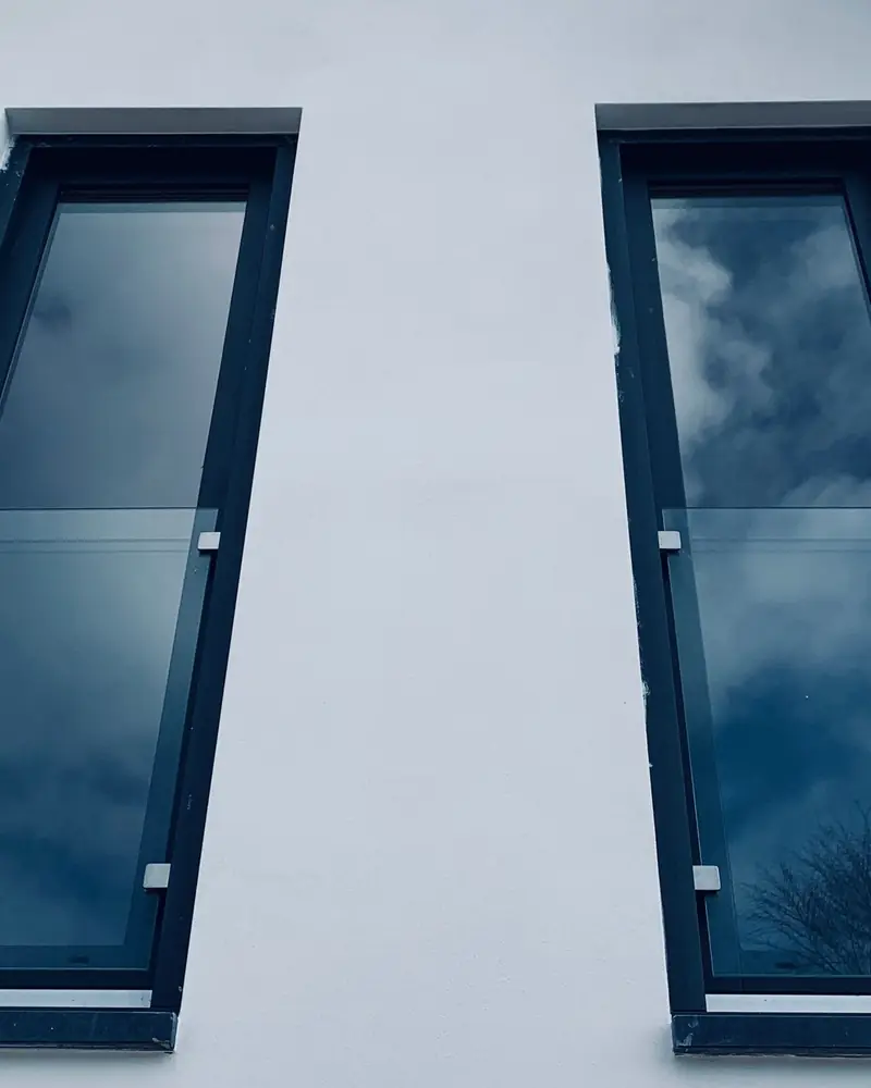 Zwei schmale bodentiefe Fenster mit dunklen Rahmen und Absturzsicherung aus Glas in einem weiß verputzten Giebel