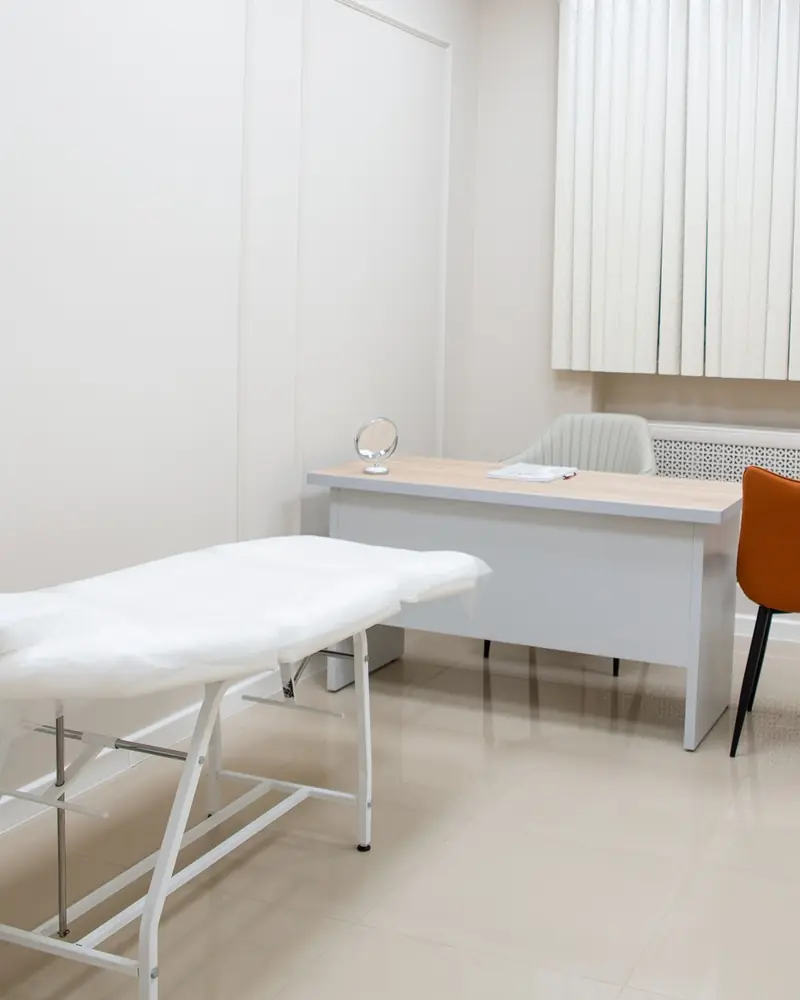 Helles Sprechzimmer mit Untersuchungsliege, Schreibtisch, zwei Stühlen und Lamellenvorhang