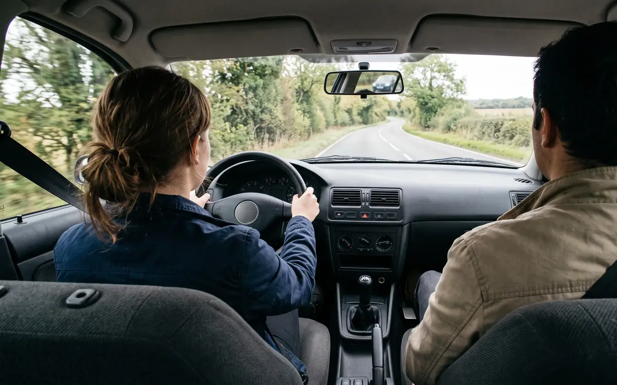 Blick von der Rückbank über die Schulter eines Fahrschülers: beide Hände am Lenkrad, rechts im Anschnitt der Fahrlehrer, vor dem Auto eine Landstraße mit Bäumen