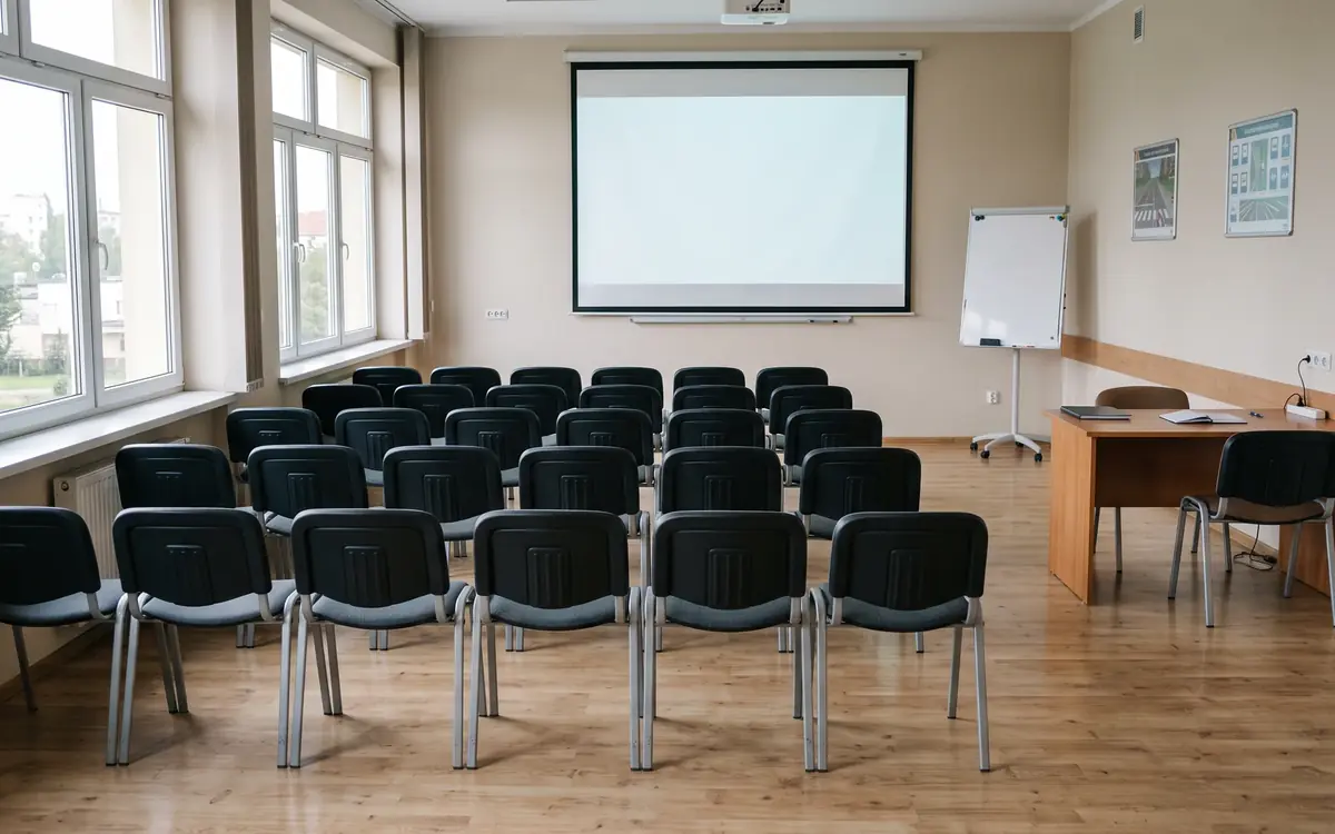 Leerer Unterrichtsraum mit Stuhlreihen vor einer Leinwand, links hohe Fenster, rechts ein Lehrertisch und ein Flipchart