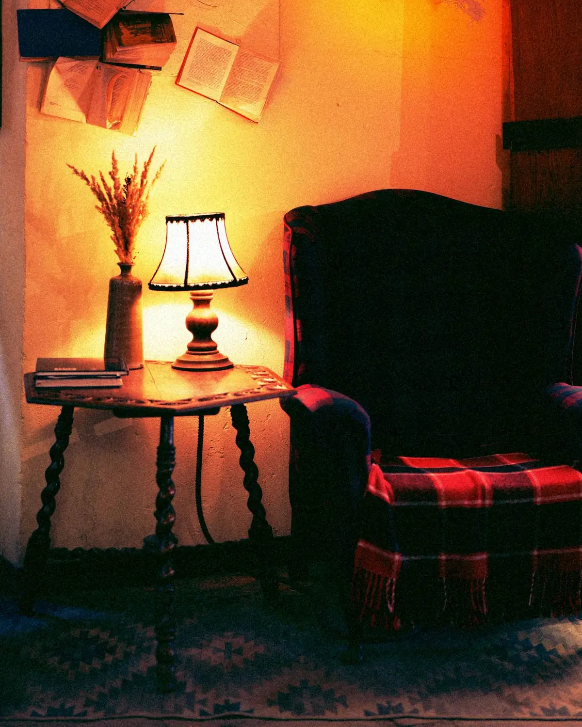 Leseecke mit Ohrensessel und karierter Decke, daneben ein gedrechseltes Tischchen mit Lampe, an der Wand aufgeschlagene Bücher