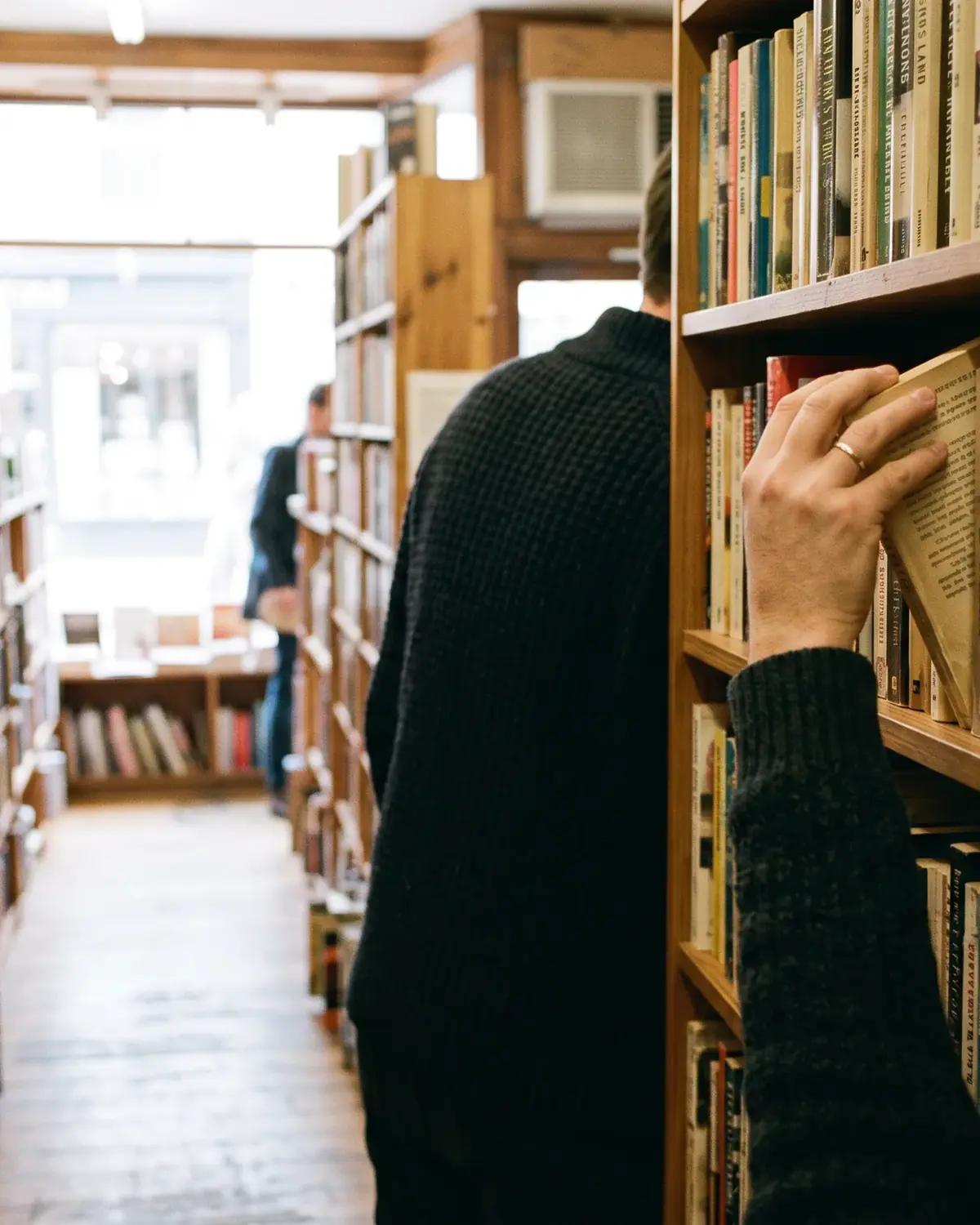Eine Person im dunklen Pullover, von hinten, zieht ein Taschenbuch aus einem langen Holzregal in einem Gang der Buchhandlung, rechts und links stehen Bücher