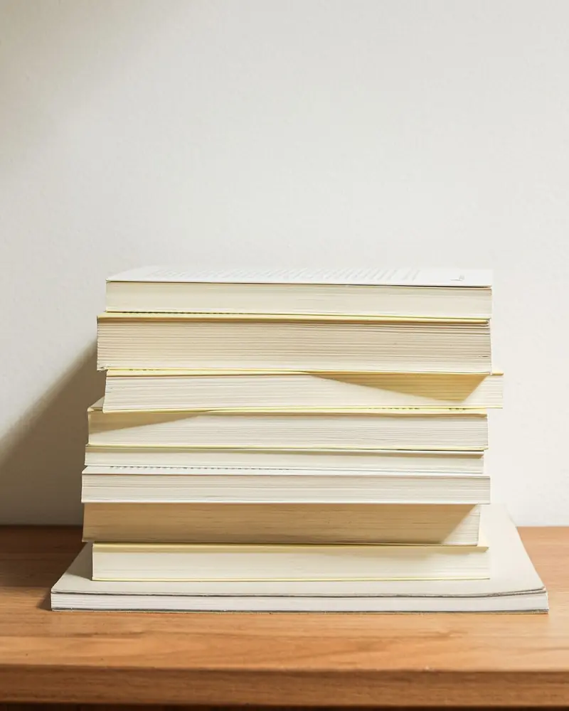 Stapel heller Taschenbücher ohne sichtbare Titel auf einem Holzbrett vor einer weißen Wand